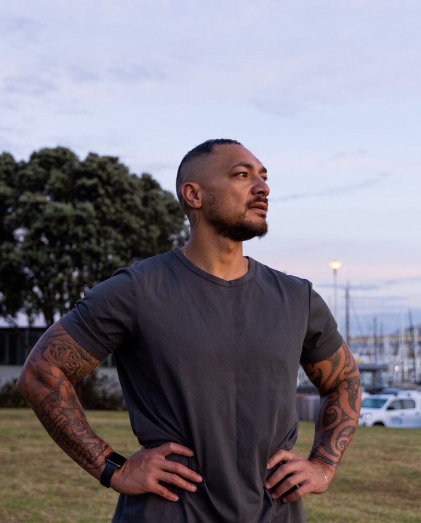 A Māori man with arm tattoos looks into the distance.